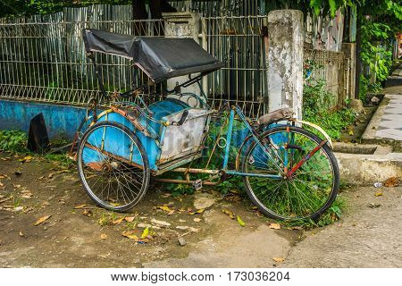 Blue pedicap or tricycle parked in side of the road near bush and wall with nobody around photo taken in Depok Indonesia java