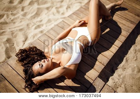 Portrait of attractive sensual woman in bikini with long wavy hair in wind with eyes closed posing on wooden path on beach with tents on background.