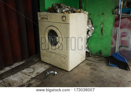 An abandoned and unused white washing machine with front door near green wall and a broom photo taken in Depok Indonesia java