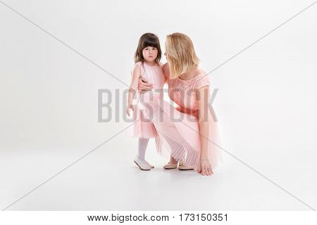 blonde mom and sweet little daughter in pink dresses princesses hugging and laughing on gray background. family