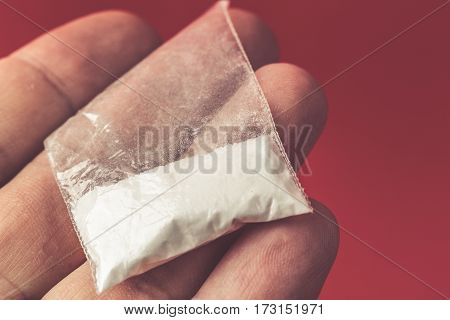 Mans hand holding or giving, selling on palm plastic packet with cocaine powder or another drugs on red background. Close-up macro retro toned photo.