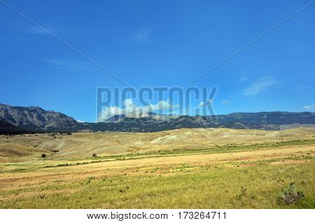 Cloud hovers over mountain in Yellowstone National Park. Vast plain and rolling hills rise at base of mountain. Blue sky is overhead.