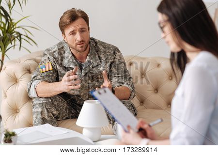 Posttraumatic stress disorder. Depressed handsome young soldier looking at his psychologist and telling her about his problems while having a posttraumatic stress disorder