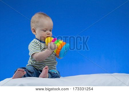 Serious little baby boy, sitting on the white blanket with a toy in hands , studio shot, isolated on blue background.