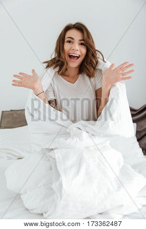 Picture of young cheerful lady dressed in white t-shirt sitting on bed at home indoors.