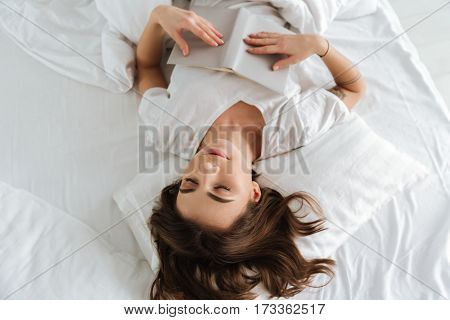 Image of young happy woman dressed in white t-shirt lies in bed at home indoors and sleeping holding book.