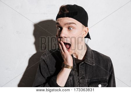 Surprised hipster in snap back holding hand on cheek with open mouth and looking away over gray background