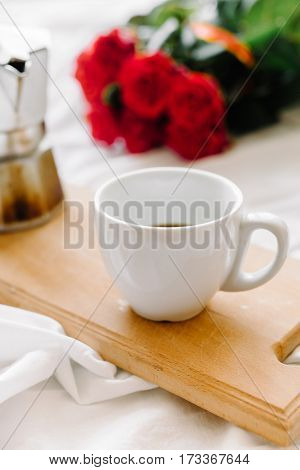 A cup of coffee coffee on the wooden Boards a bouquet of red roses on a white rumpled bedding greeting early in the morning