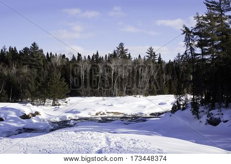 Wide view of a snowy country landscape with a small stream of rapids running through and a background of green fir trees at Pabineau Falls near Bathurst New Brunswick on a bright sunny day with blue skies and clouds in February.