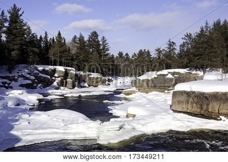 Wide view of a snowy country landscape with a small stream of rapids running through rock faces on either side and a background of green fir trees at Pabineau Falls near Bathurst, New Brunswick on a bright sunny day with blue skies and clouds in February.