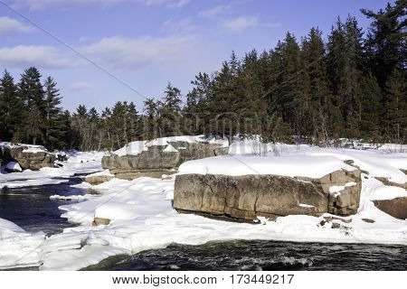 Wide view of a snowy country landscape with a small stream of rapids running through rock faces on either side and a background of green fir trees at Pabineau Falls near Bathurst New Brunswick on a bright sunny day with blue skies and clouds in February.