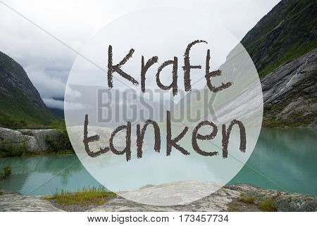 German Text Kraft Tanke Means Relax. Lake With Mountains In Norway. Cloudy Sky. Peaceful Scenery, Landscape With Rocks And Grass. Greeting Card