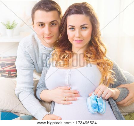 Happy Young Couple family Expecting Baby. Beautiful Pregnant Woman and Her Husband Together Caressing Her Pregnant Belly at Home on a Sofa. Expectant parents, Mom and Dad waiting for their baby.