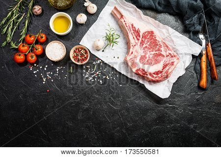 Raw tomahawk beef steak with ingredients for grilling: seasoning, fresh rosemary and olive oil on paper over black background, top view