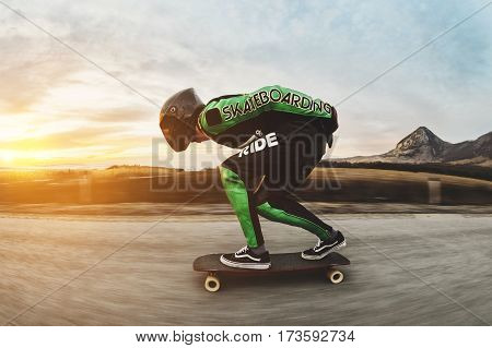 A man in a fullface helmet and a leather suit in a special rack rides a longboard on afsaltu sunset in the background mountains and beautiful sky at high speed.