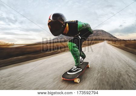 A man in a fullface helmet and a leather suit in a special rack rides a longboard on afsaltu sunset in the background mountains and beautiful sky at high speed.