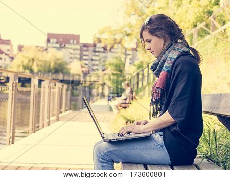 Young woman using laptop at a riverbank. Milleanial working in a park.