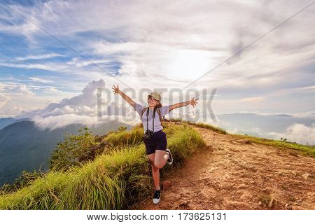Happy hiker cute teen girl with dslr camera cap eyeglass and backpack stand smiling open arms on peak mountain and sky during sunset background at Phu Chi Fa Forest Park Chiang Rai Thailand