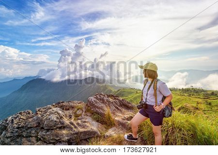 Hiker asian teen girl happy with camera backpack cap and eyeglass looking beautiful landscape nature of mountain range and colourful sky at sunset on Phu Chi Fa Forest Park Chiang Rai Thailand
