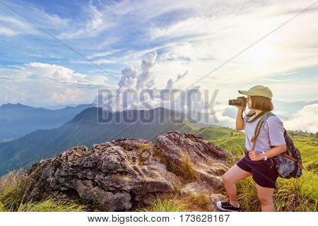 Hiker asian teen girl wear cap and eyeglass with happy holding dslr camera shooting photo beautiful nature in winter on peak mountain at sunset in Phu Chi Fa Forest Park Chiang Rai Thailand