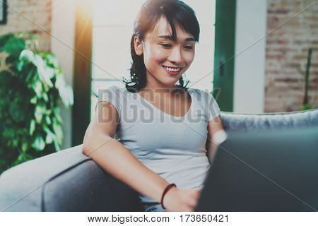 Portrait of attractive young Asian woman sitting at sofa and working modern laptop at home, wearing white tshirt.Horizontal, blurred backgroung, flare effect