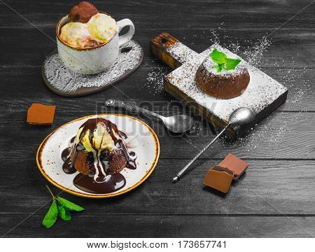 Chocolate lava cake (Molten) with ice cream on plate. Balls of ice cream in cup. Chocolate lava cake (Molten) with mint on board. Pieces of chocolate. Dark black wooden background.