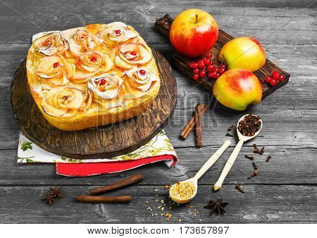 Fruit apple pie tartlet (cake). On top of the pie cake in shape of apples rose flowers. Fresh fruit apples for pie cake. Spice cloves star anise berries orange peel. Gray wood background.