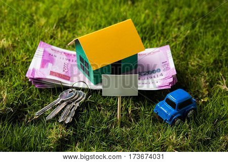 indian currency notes of rupees 100, 500 and 2000 in roll or stack with small home model, keys, toy car and calculator with blank board over green grass - home buying and finance concept in India