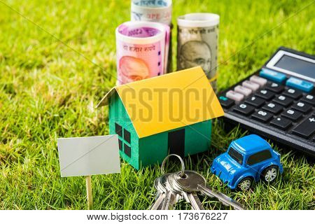 indian currency notes of rupees 100, 500 and 2000 in roll or stack with small home model, keys, toy car and calculator with blank board over green grass - home buying and finance concept in India
