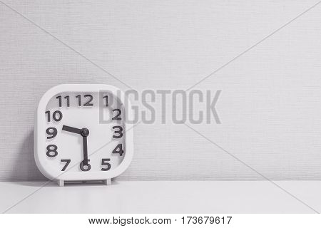 Closeup white clock for decorate show half past nine or 9:30 a.m. on white wood desk and cream wallpaper textured background in black and white tone with copy space