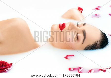 Beautiful Fashion model girl in milk bath, perfect face skin, spa and skin care concept. Beauty young Woman with red rose flower relaxing in milk bath. Healthy spa procedures, rejuvenation