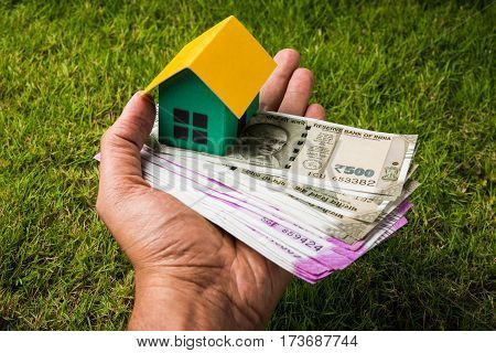 hand holding new indian currency notes or rupees 500 and 2000 and house model over green grass - indian real estate and finance concept