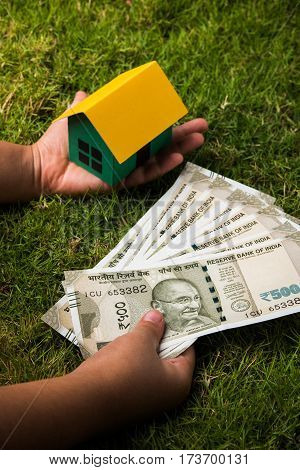 hand holding new indian currency notes or rupees 500 and 2000 and house model over green grass - indian real estate and finance concept