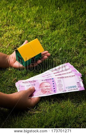 hand holding new indian currency notes or rupees 500 and 2000 and house model over green grass - indian real estate and finance concept