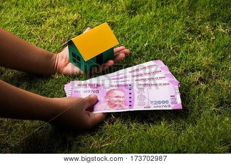 hand holding new indian currency notes or rupees 500 and 2000 and house model over green grass - indian real estate and finance concept