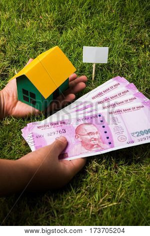 hand holding new indian currency notes or rupees 500 and 2000 and house model over green grass - indian real estate and finance concept