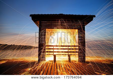 Hot Golden Sparks Flying from Man Spinning Burning Steel Wool on Wooden Bridge Extended into the Sea. Long Exposure Photography using Steel Wool Burning.