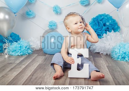 Portrait of cute adorable Caucasian baby boy with blue eyes barefoot in pants with suspenders and hat sitting on wooden floor in studio holding large letter E looking away first year concept