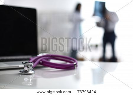 Laptop and stethoscope on the desk doctors standing in the background.