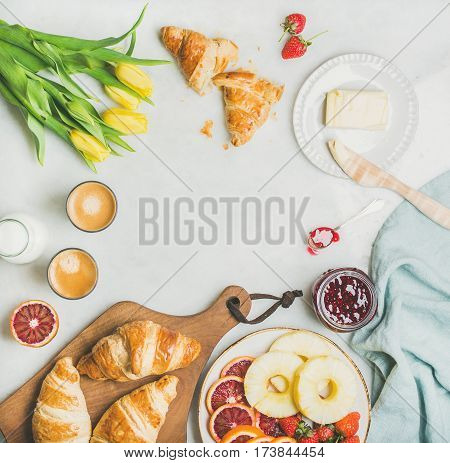 Breakfast table with croissants, butter, raspberry jam, fresh fruit, milk in bottle, coffee espresso and spring yellow tulip flowers. Top view, copy space