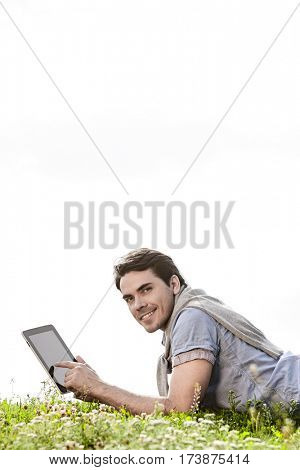 Side view portrait of young man using digital tablet while lying on grass against clear sky