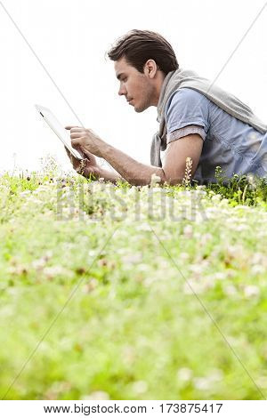 Side view of young man using digital tablet while lying on grass against clear sky