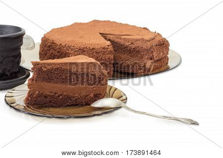 Piece of the chocolate layered cake sprinkled with cocoa powder and the spoon on a dark glass saucer on the background of the rest of the cake and coffee cup closeup on a light background