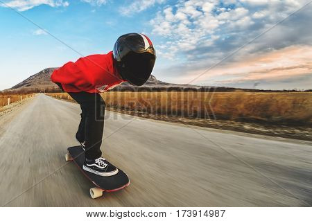 A young man in a fullface helmet and a leather suit in a special rack rides a longboard on afsaltu sunset in the background mountains and beautiful sky at high speed.