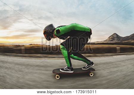 A young man in a fullface helmet and a leather suit in a special rack rides a longboard on afsaltu sunset in the background mountains and beautiful sky at high speed.