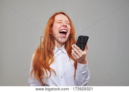 Close-up of red hair girl laughing while holding a mobile phone. Heartily laughing girl isolated over background in the studio 