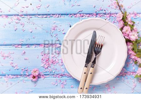 Springr table setting. Pink flowers knife and fork on white plate on blue wooden background. Selective focus. Place for text. Top view.