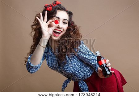 Happy Beautiful Pin Up Girl Holding Cup of Glass Bottle as OK Symbol and Giving you a Soda. Woman Dressed in Red Skirt and Blue Plaid Shirt.