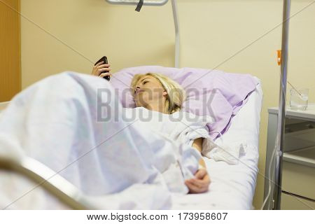 Bedridden female patient lying in hospital bed, using mobile phone while recovering after surgery.