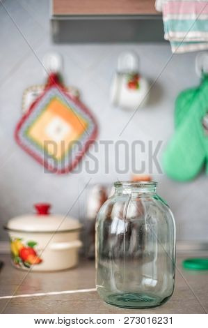 Empty Three-liter Jar Is On The Tabletop Against The Background Of The Pot And Oven Mitts.